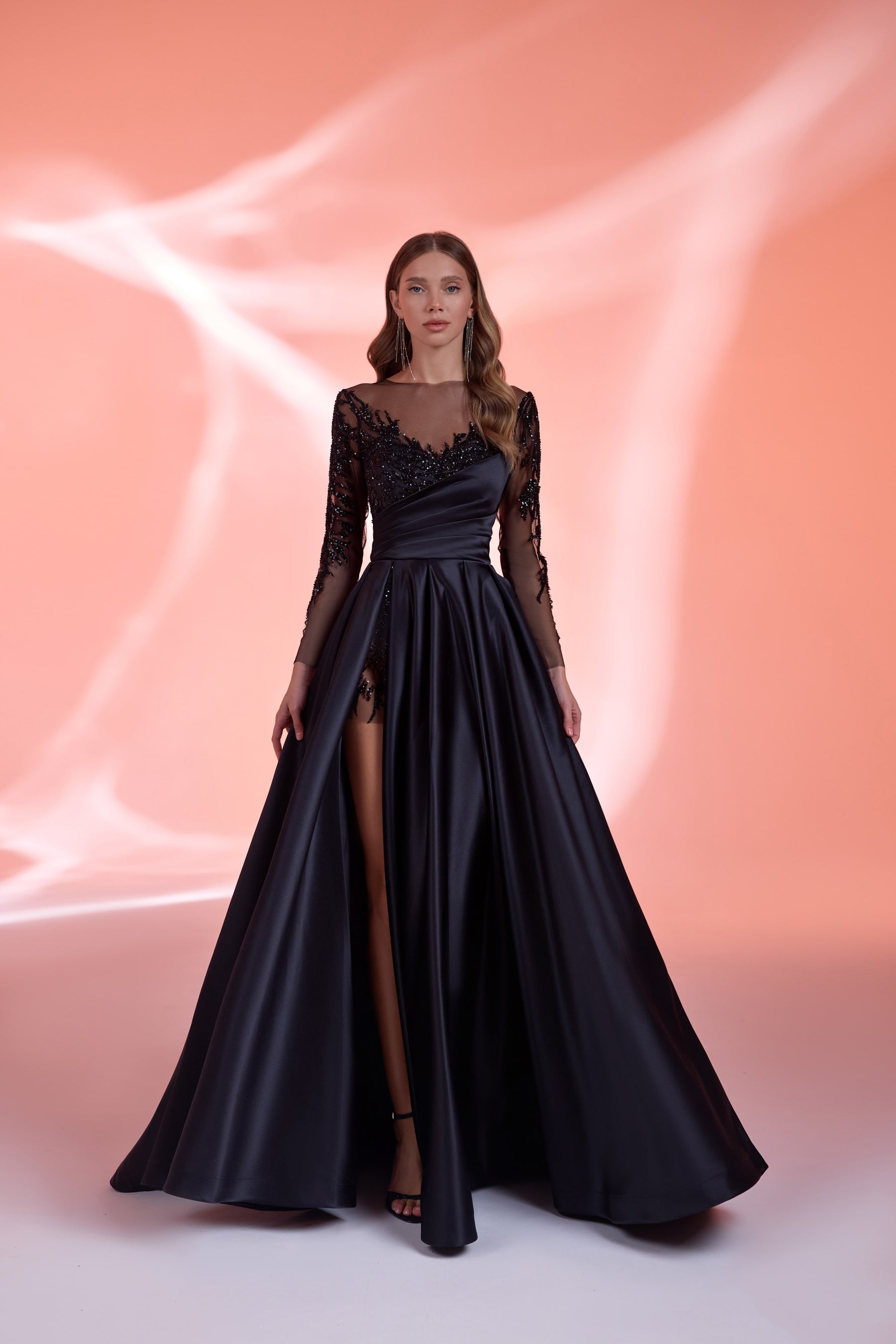 Black evening dress with a draped fitted bodice, sheer embellished sleeves, thigh-high slit, and full skirt