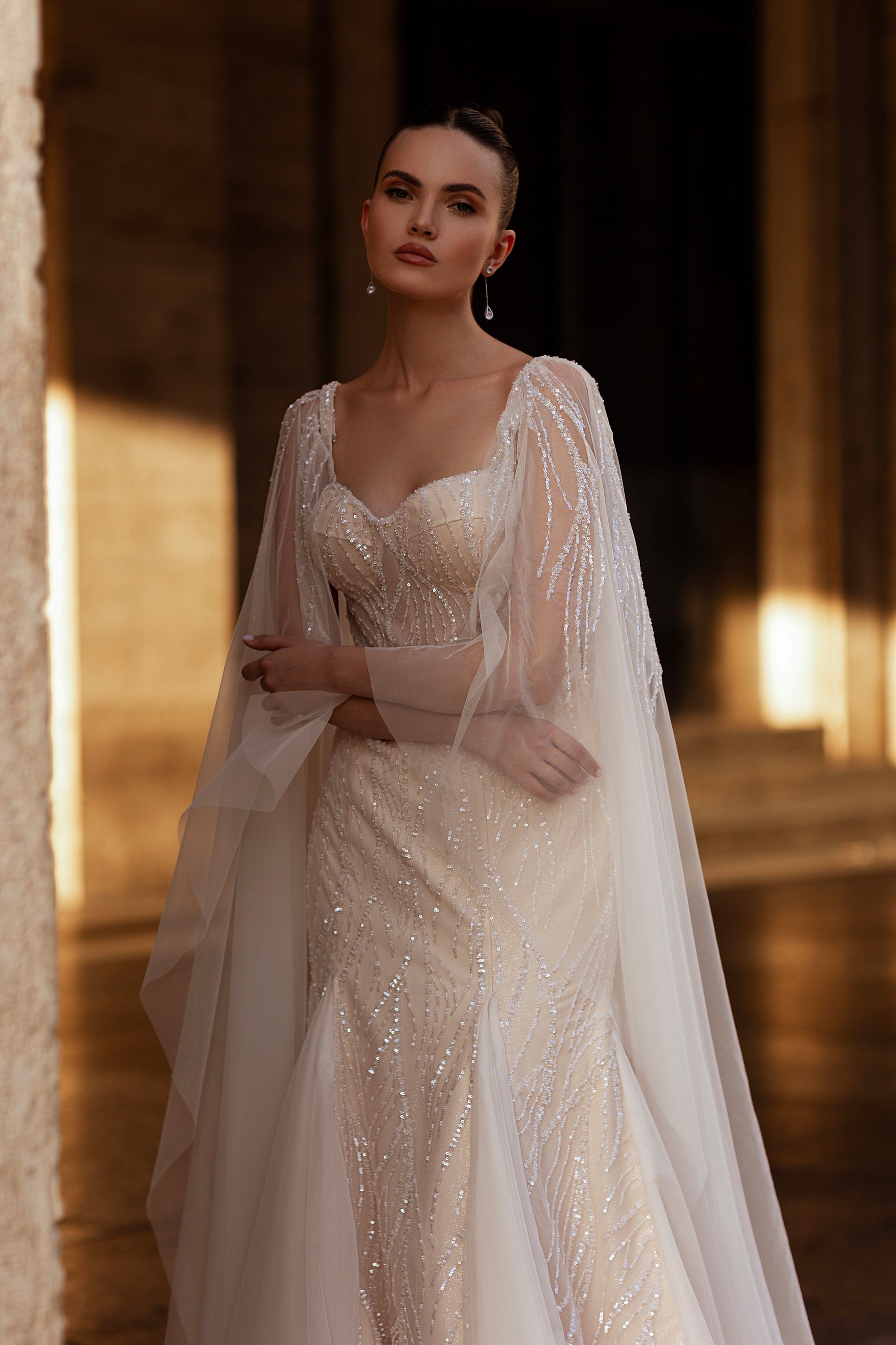 Sparkling ivory wedding dress with a sheer square neckline, beaded bodice, and draped cape