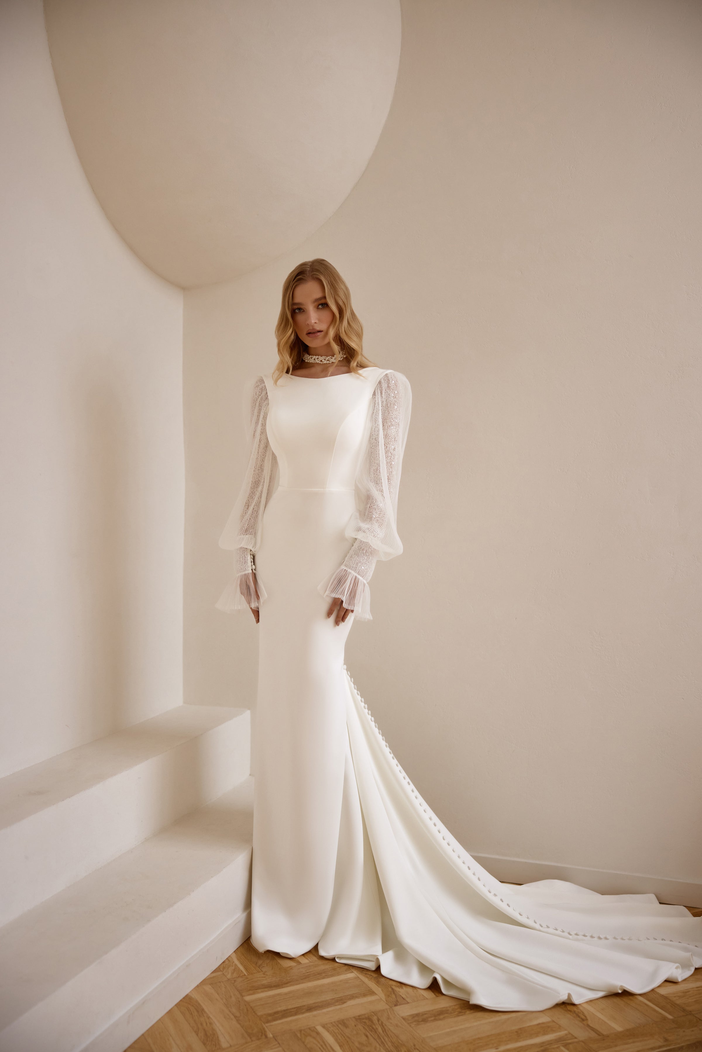 Ivory sheath wedding dress with a boat neckline, sheer lace bishop sleeves, and a long flowing train