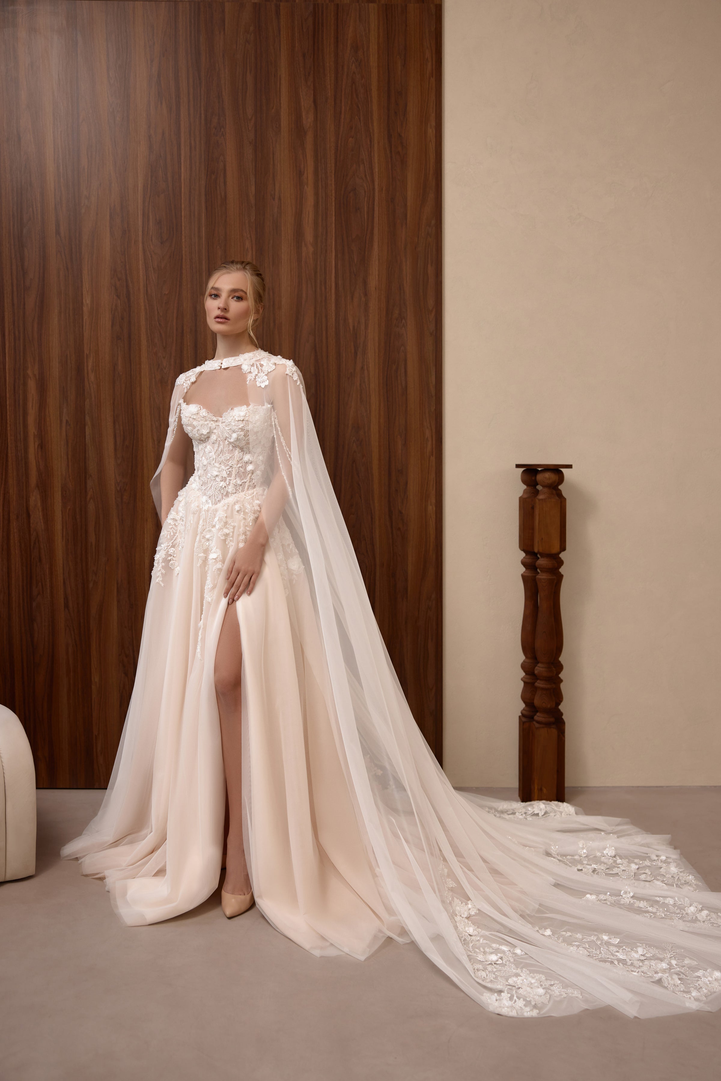 Blush A-line wedding dress with floral lace corset, sheer cape, thigh-high slit, and sweeping embellished train