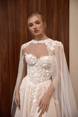 Close-up of a blush wedding dress with sheer high neckline, floral lace corset, and flowing cape