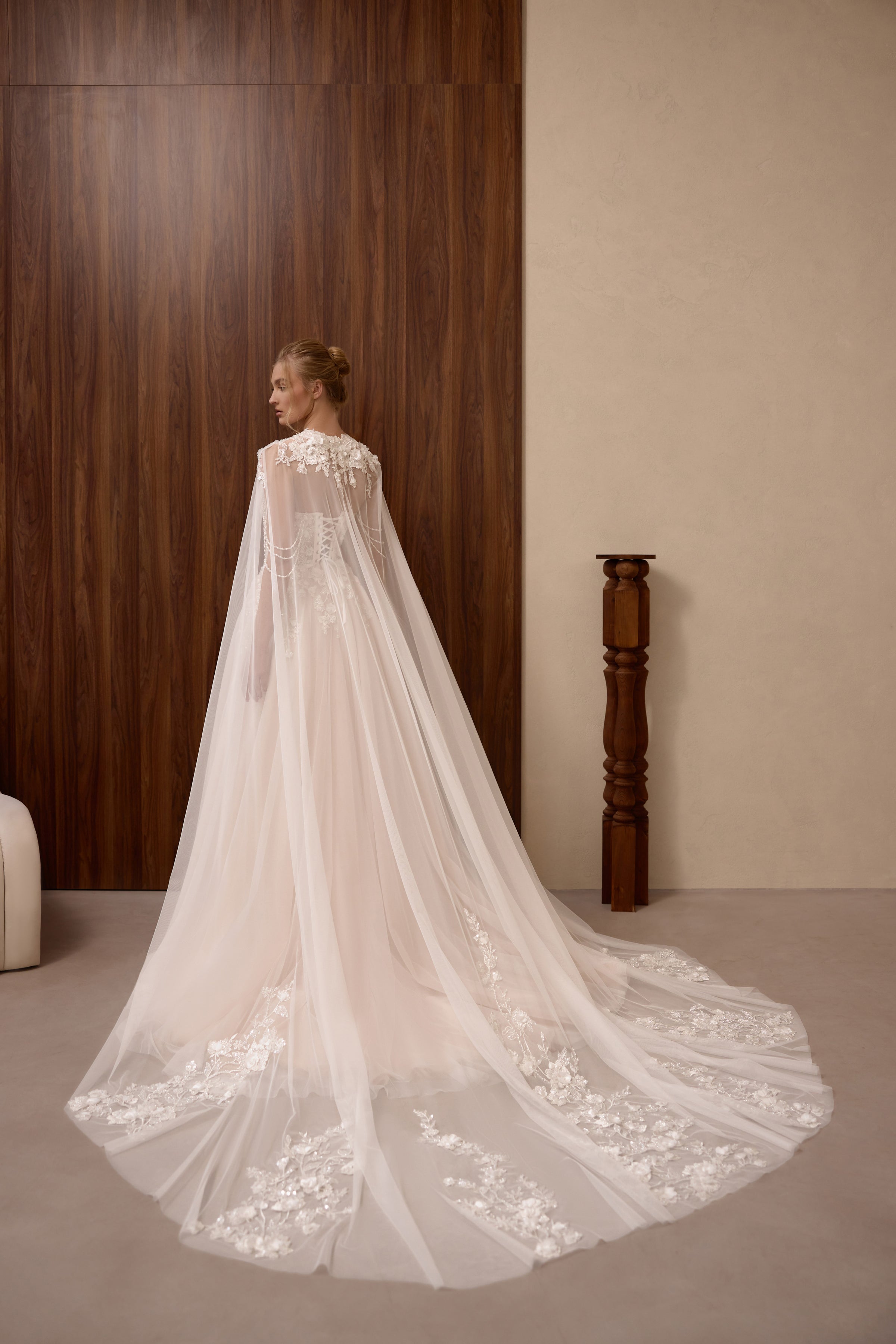 Back view of a blush wedding dress beneath a sheer floral-hem cape with a long, flowing train