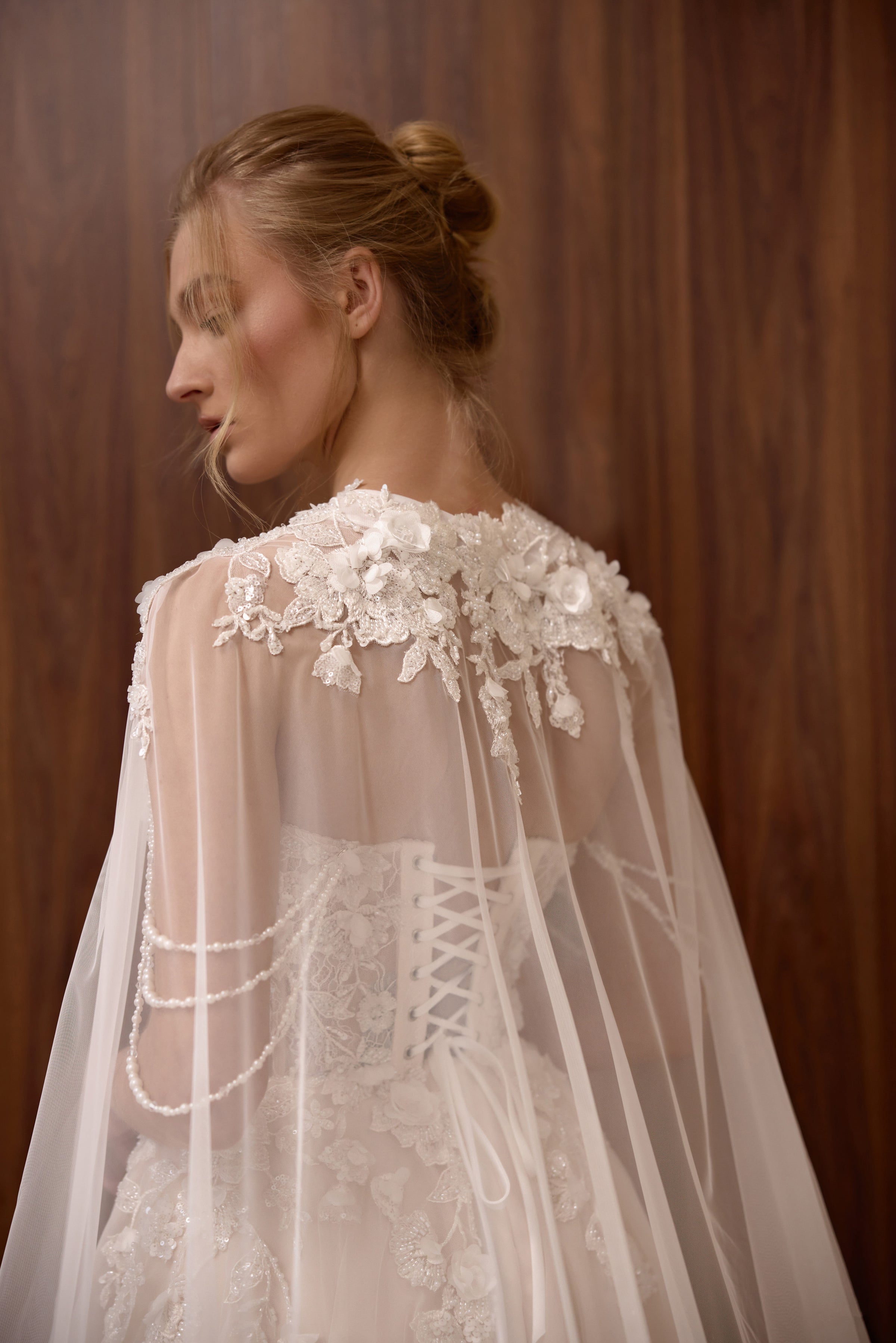Lace-up corset back and floral shoulder appliqués beneath a sheer cape on a blush wedding dress