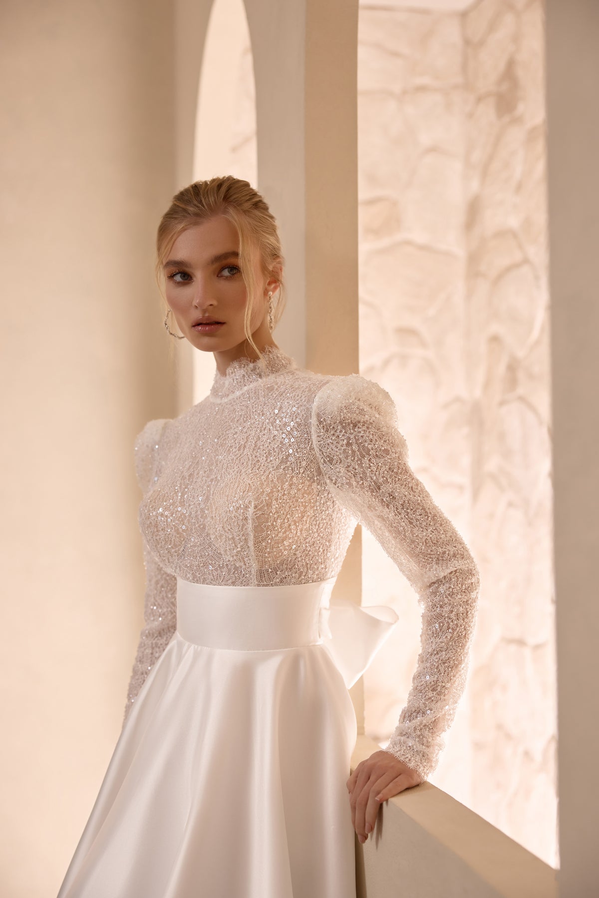 Ivory wedding dress featuring a high neckline, sparkling sheer long sleeves, fitted beaded bodice, and satin waistband