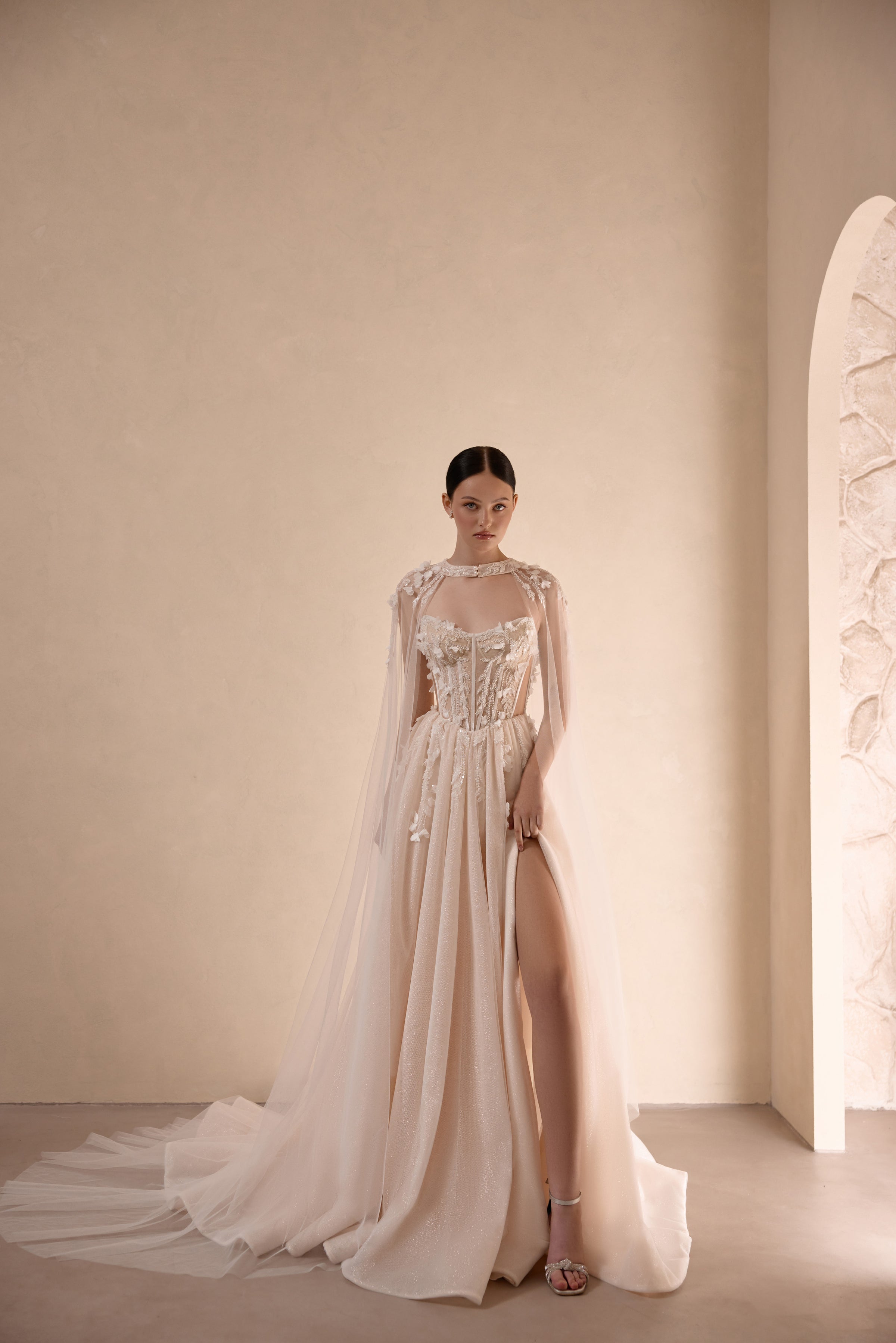 Ivory wedding dress with a beaded corset bodice, high slit, and sheer flowing cape