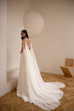 Open V-back ivory wedding dress with a flowing cape and sweeping train