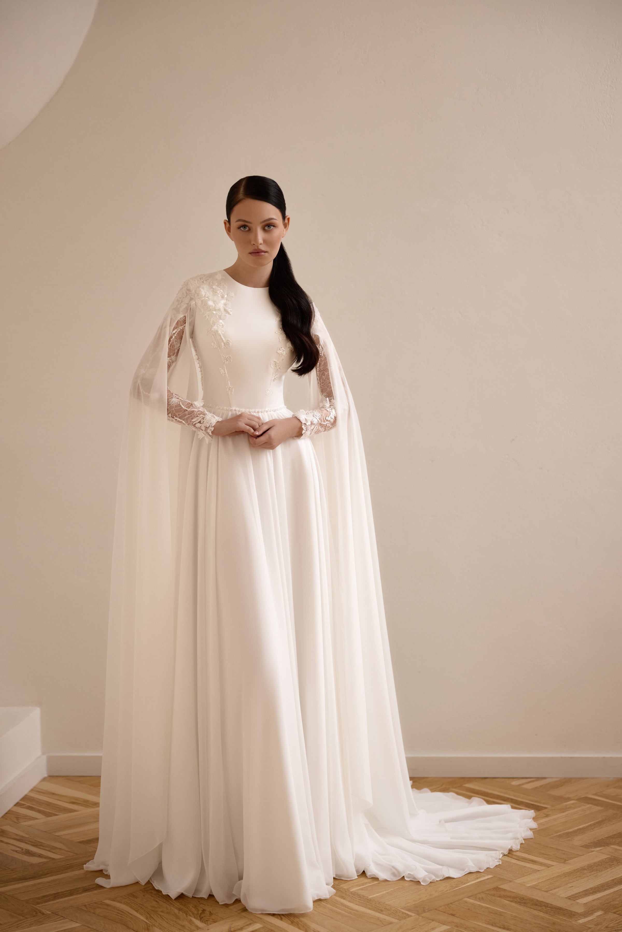 Ivory A-line wedding dress with a high neckline, sheer lace sleeves, flowing cape, and long train