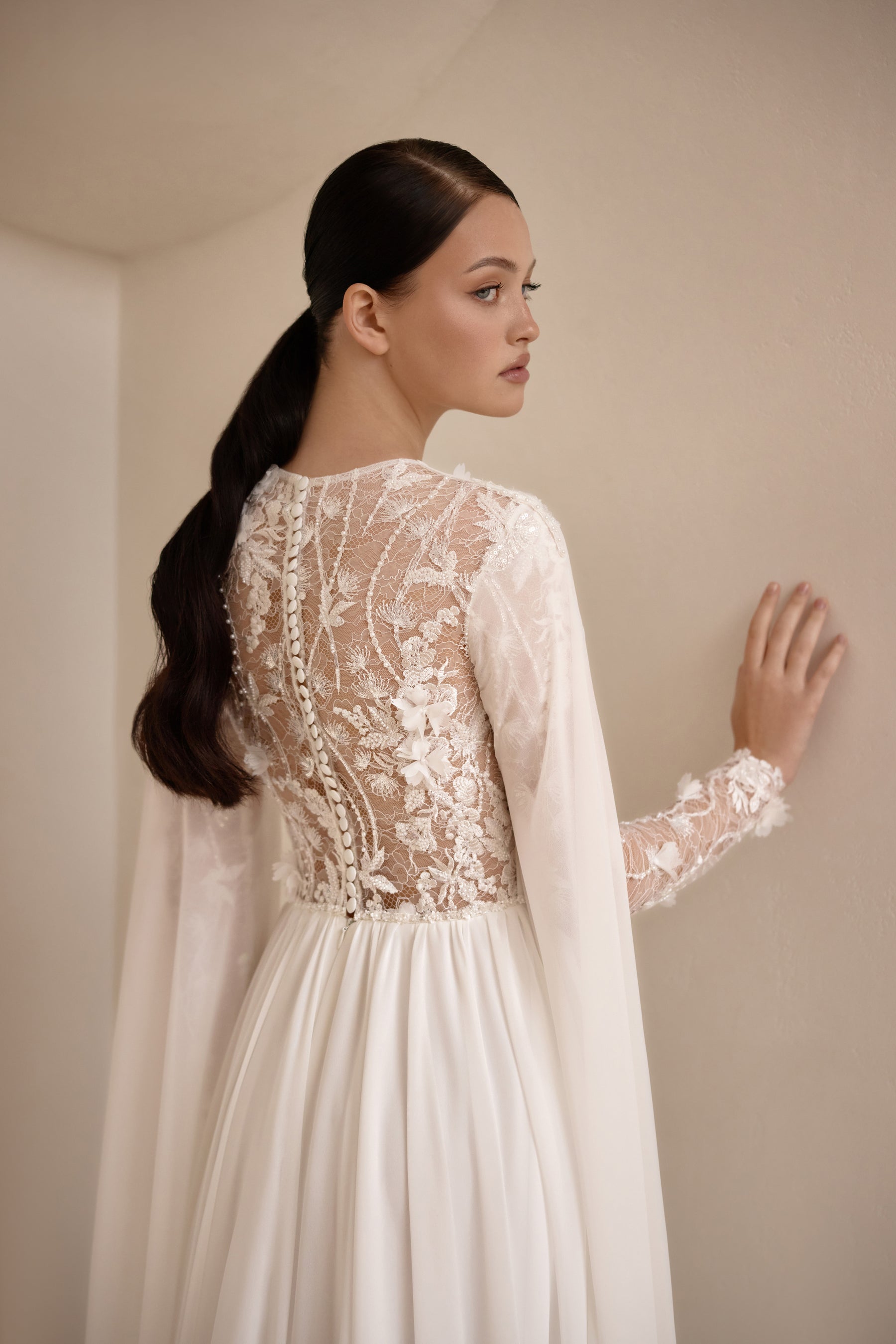 Close back detail of an ivory wedding dress with floral lace overlay, sheer sleeves, and flowing cape