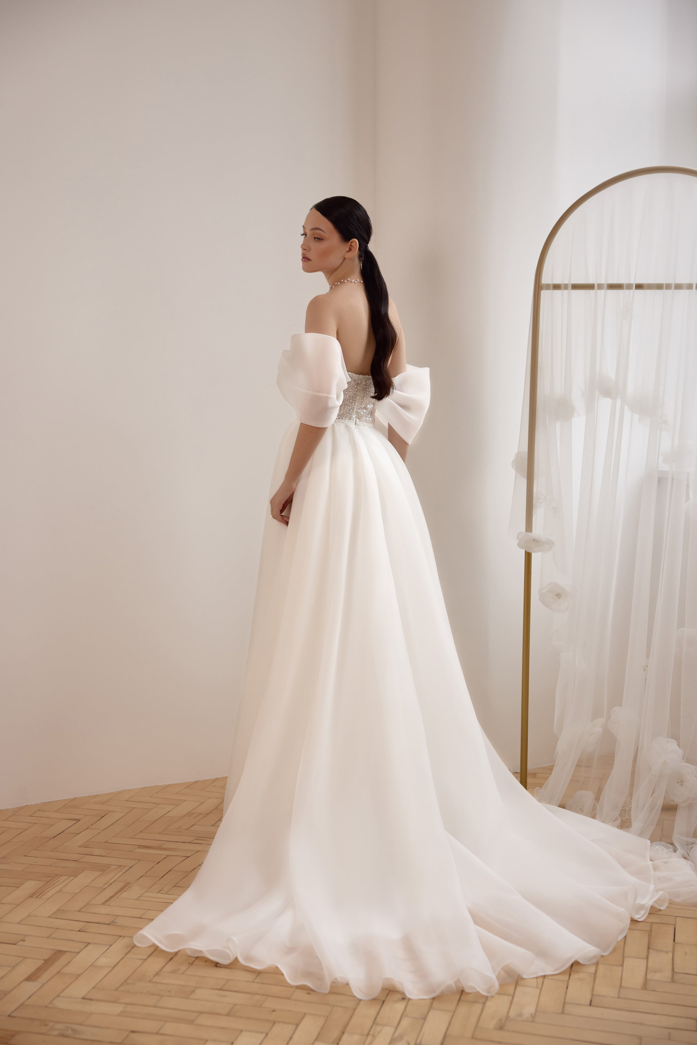 Three-quarter back view of an ivory wedding dress with off-the-shoulder sleeves, a large waist bow, and long train