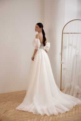 Three-quarter back view of an ivory wedding dress with off-the-shoulder sleeves, a large waist bow, and long train