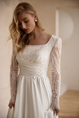 Close-up of an ivory wedding dress bodice with a square neckline, shimmering lace, and sheer long sleeves