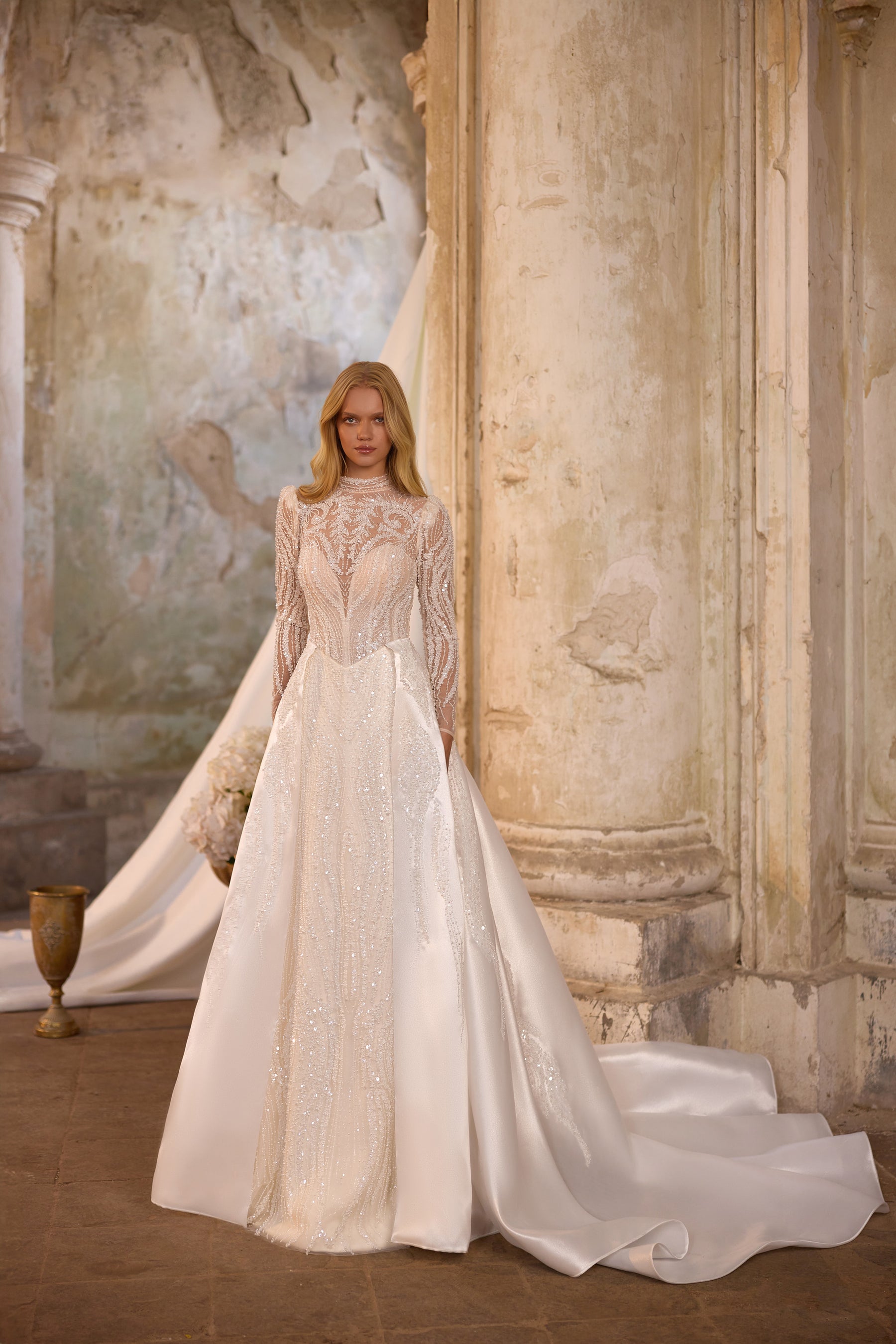 Ivory wedding dress shown with a detachable full overskirt, long lace sleeves, and a beaded high neckline