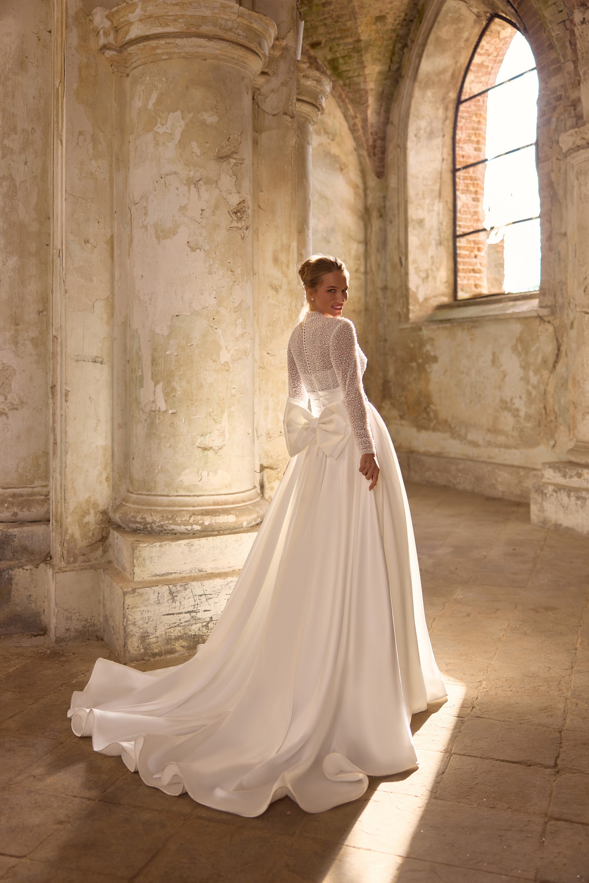 Ivory wedding dress with a sheer beaded high-neck back, long sleeves, waist bow, and flowing train