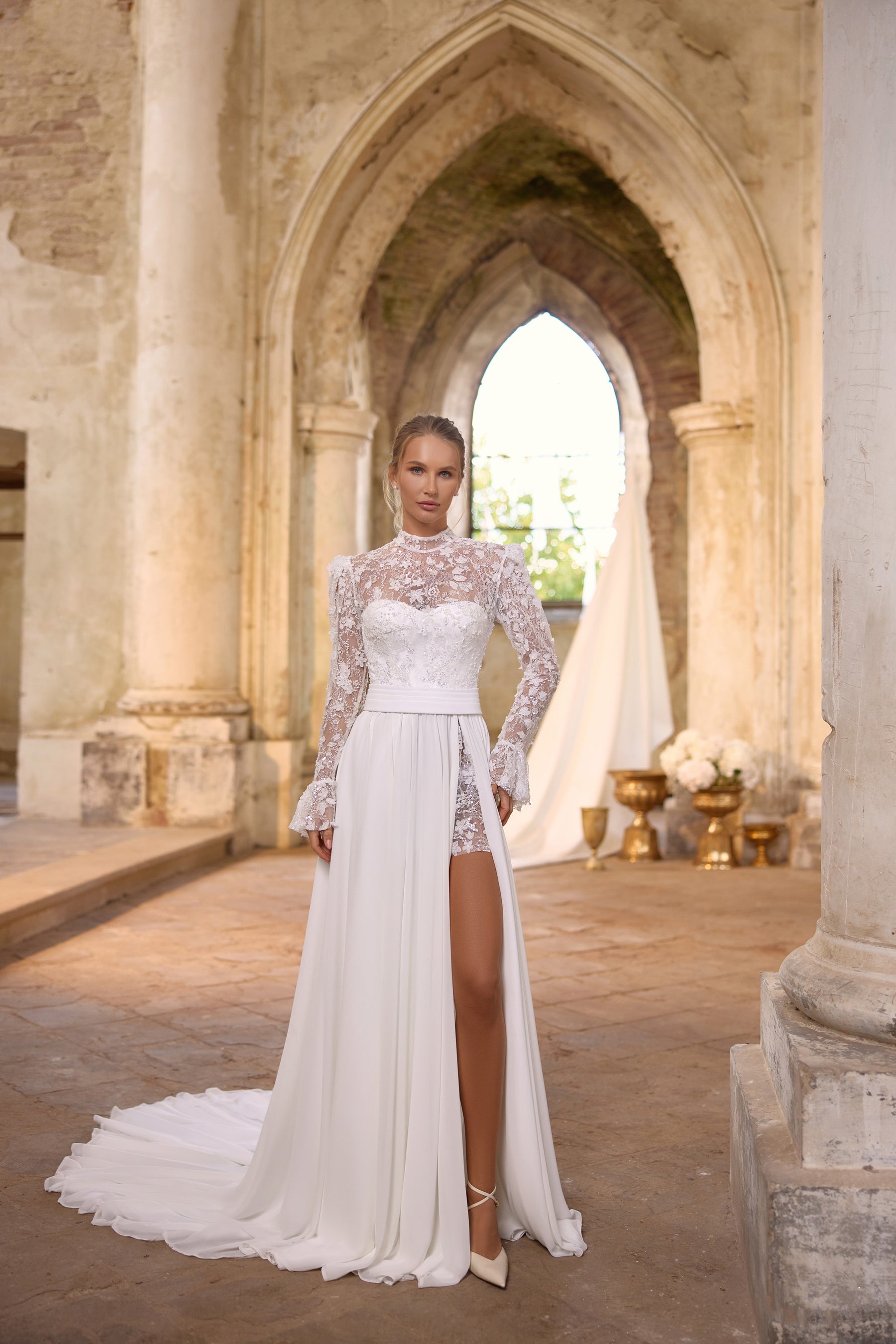 Ivory wedding dress with a high lace neckline, long sleeves, waist sash, and flowing slit overskirt