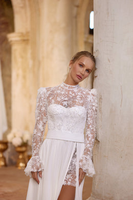 Close-up of an ivory wedding dress with a sheer floral lace bodice, high neckline, flared cuffs, and slit