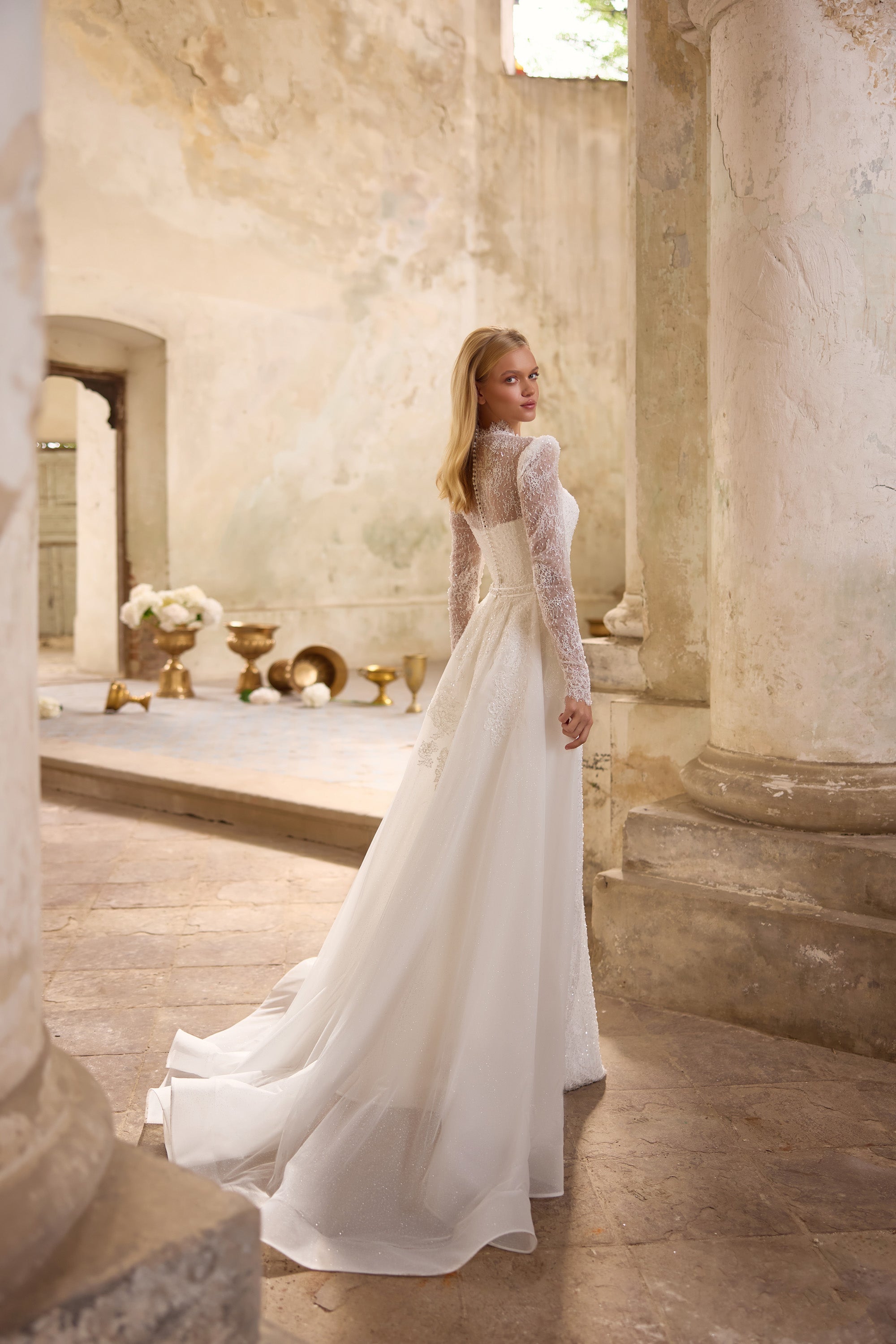Ivory wedding dress showing a sheer lace back, long sleeves, flowing overskirt, and extended train