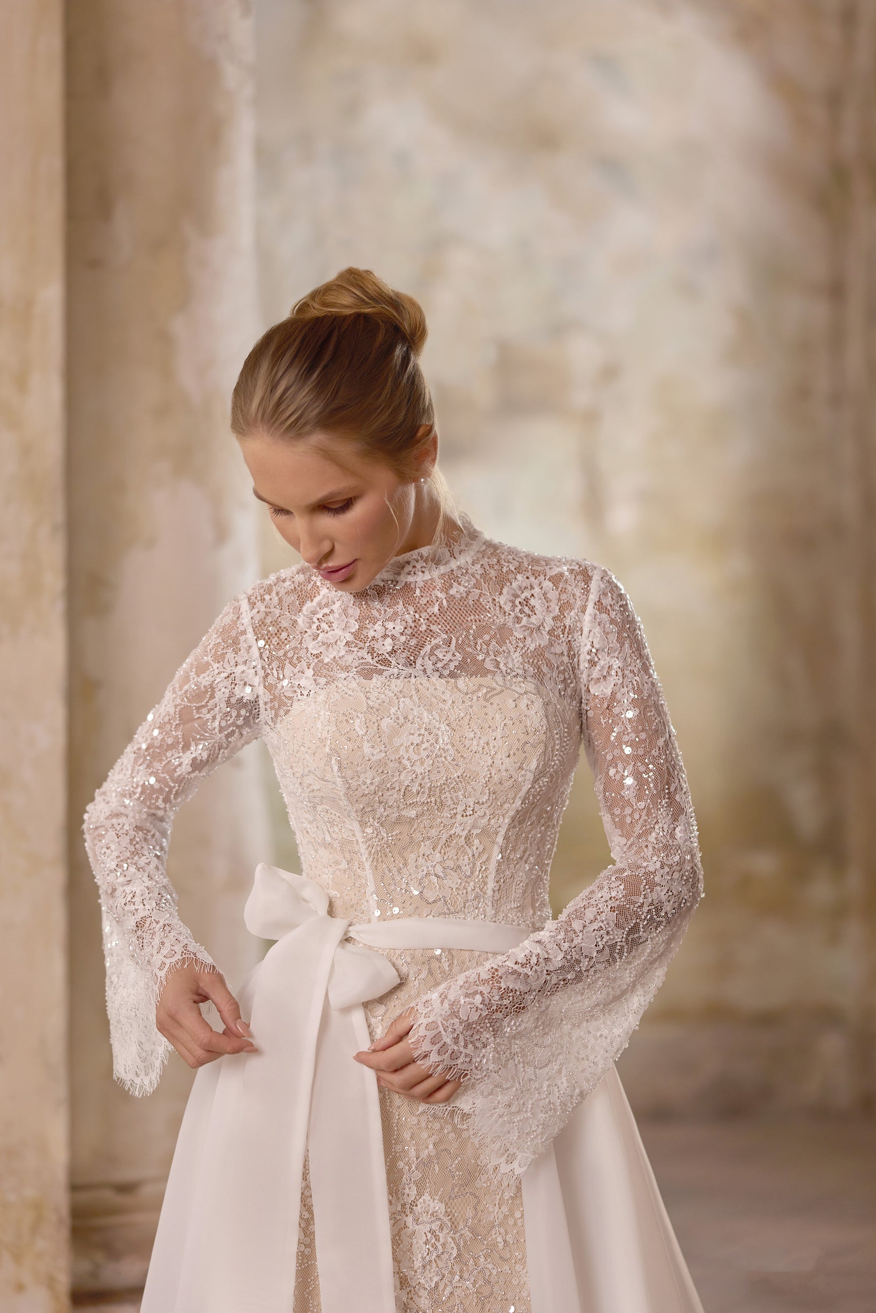 Close-up of an ivory wedding dress with a high lace neckline, sheer long sleeves, jeweled bodice, and waist bow