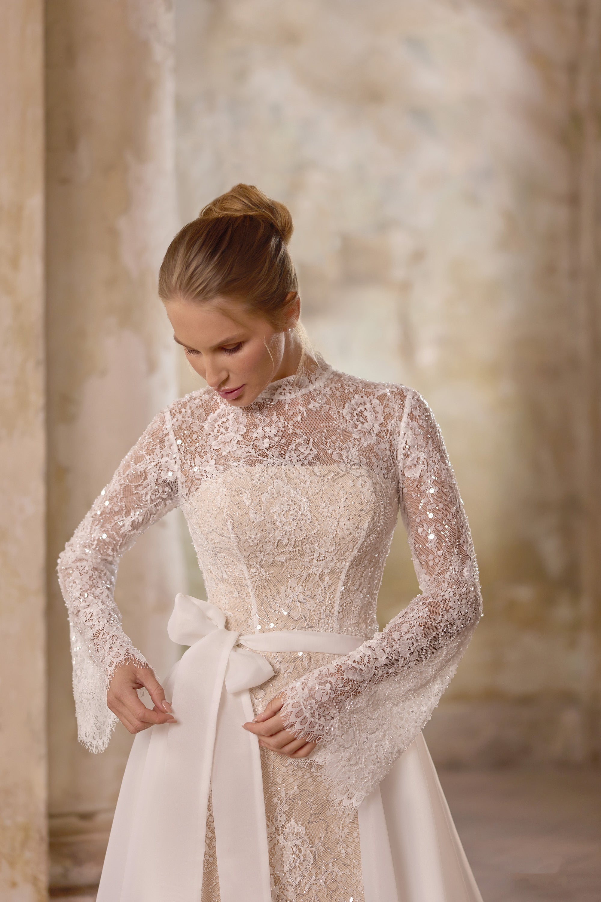 Close-up of an ivory wedding dress with a high lace neckline, sheer long sleeves, jeweled bodice, and waist bow