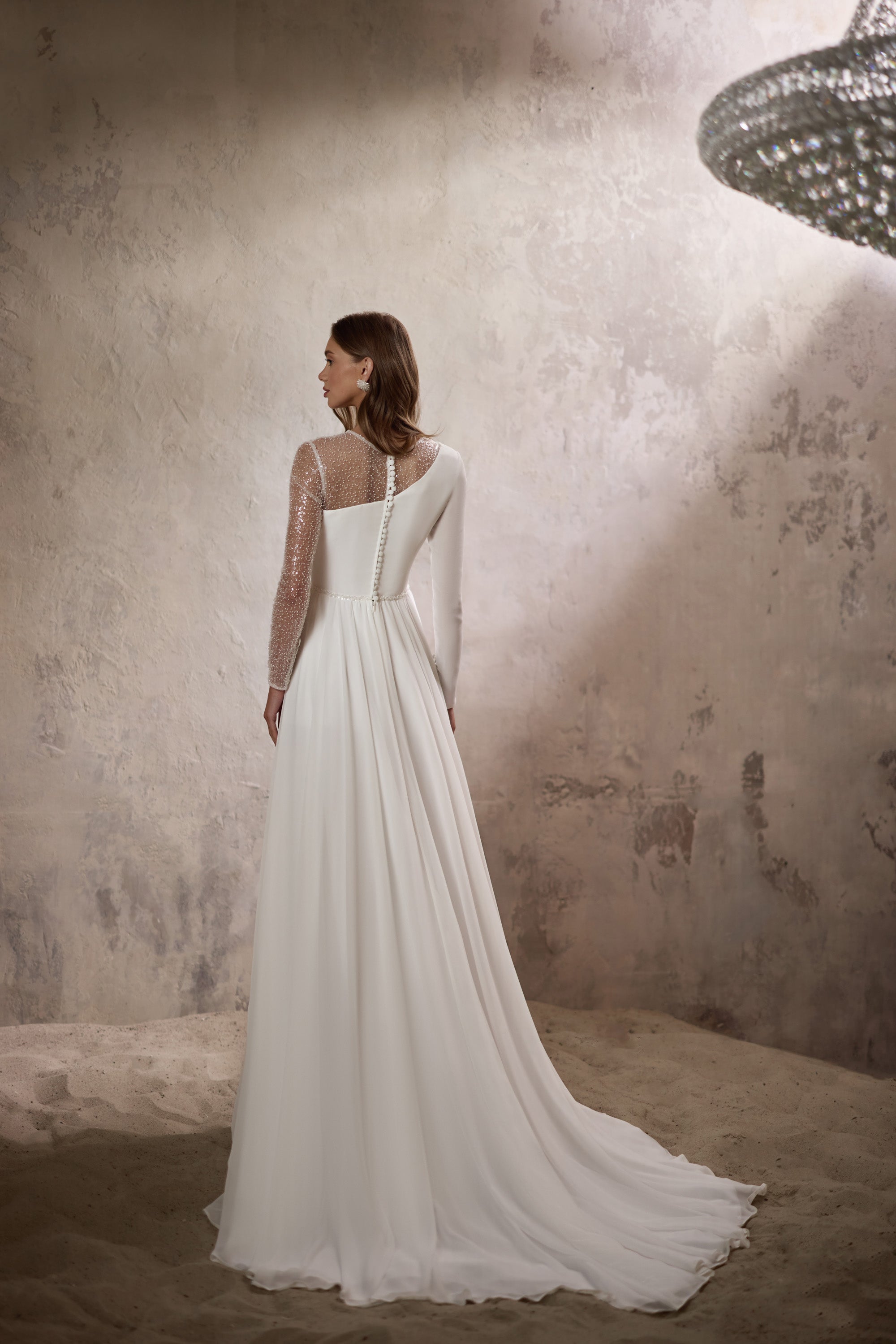 Back of an ivory wedding dress with a sheer sequined sleeve, defined waist, flowing skirt, and long train