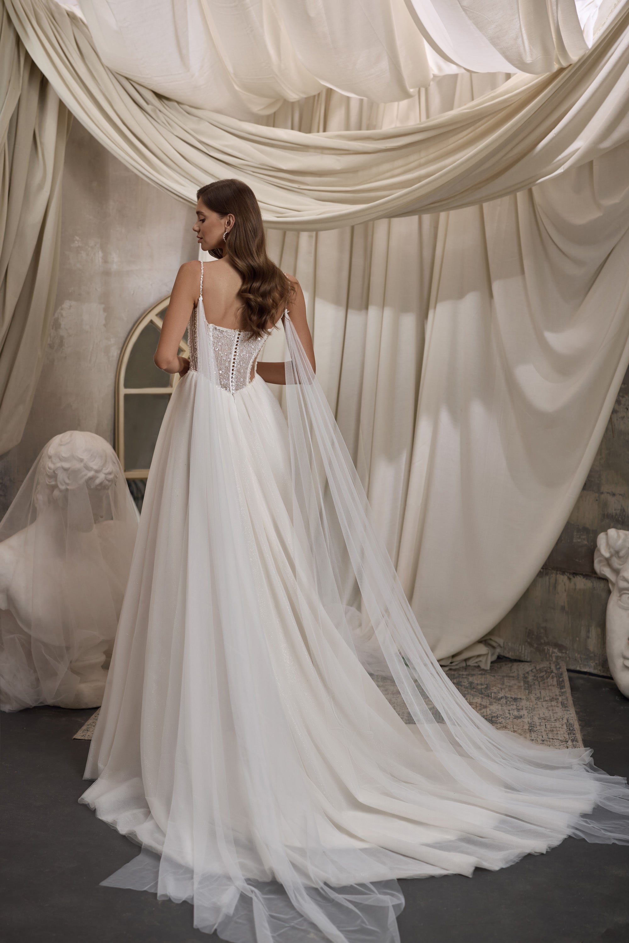Back view of an ivory wedding dress with an embellished open back, thin straps, and sheer trailing cape