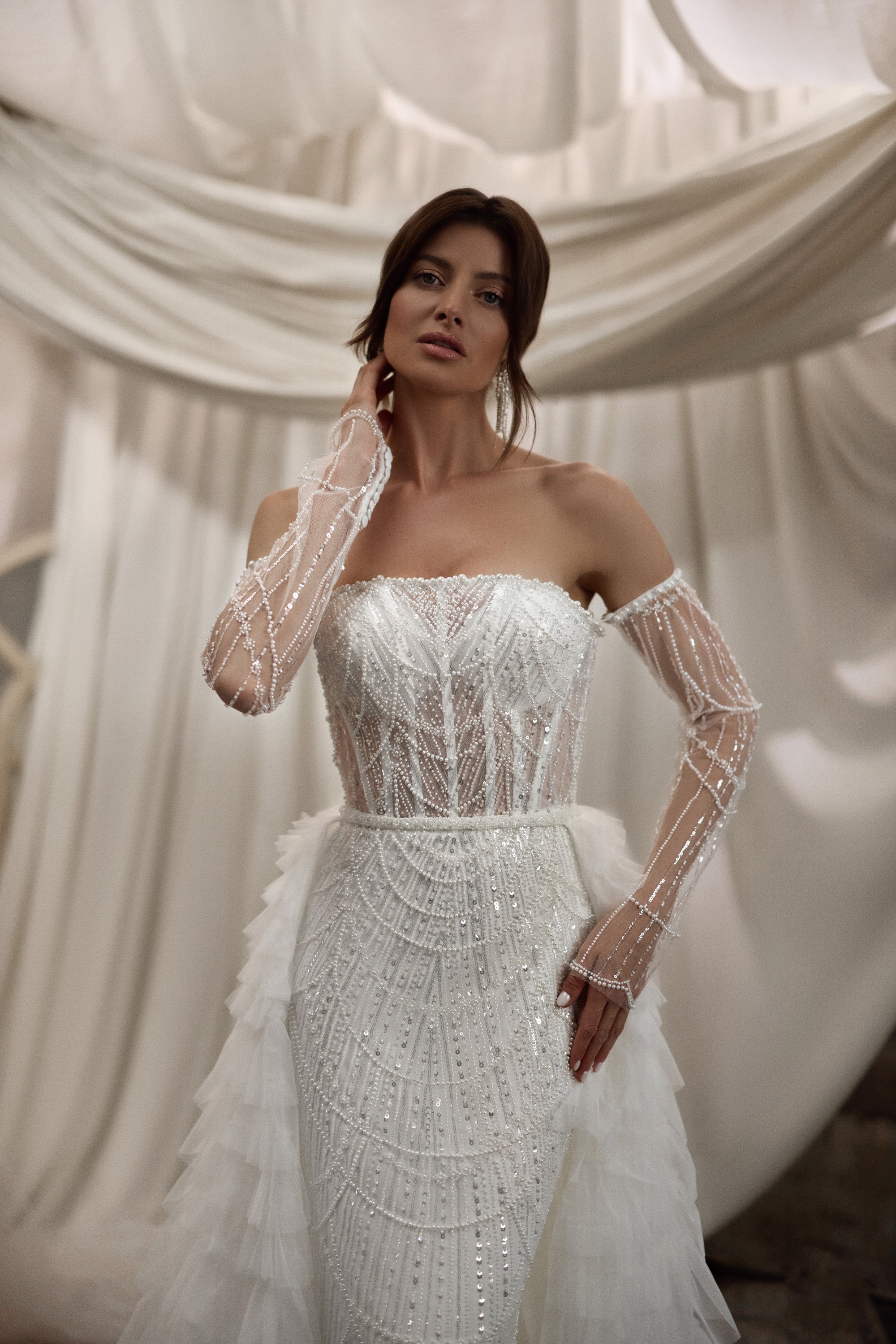 Beaded ivory wedding dress with a strapless bodice, sheer embellished sleeves, and ruffle overskirt