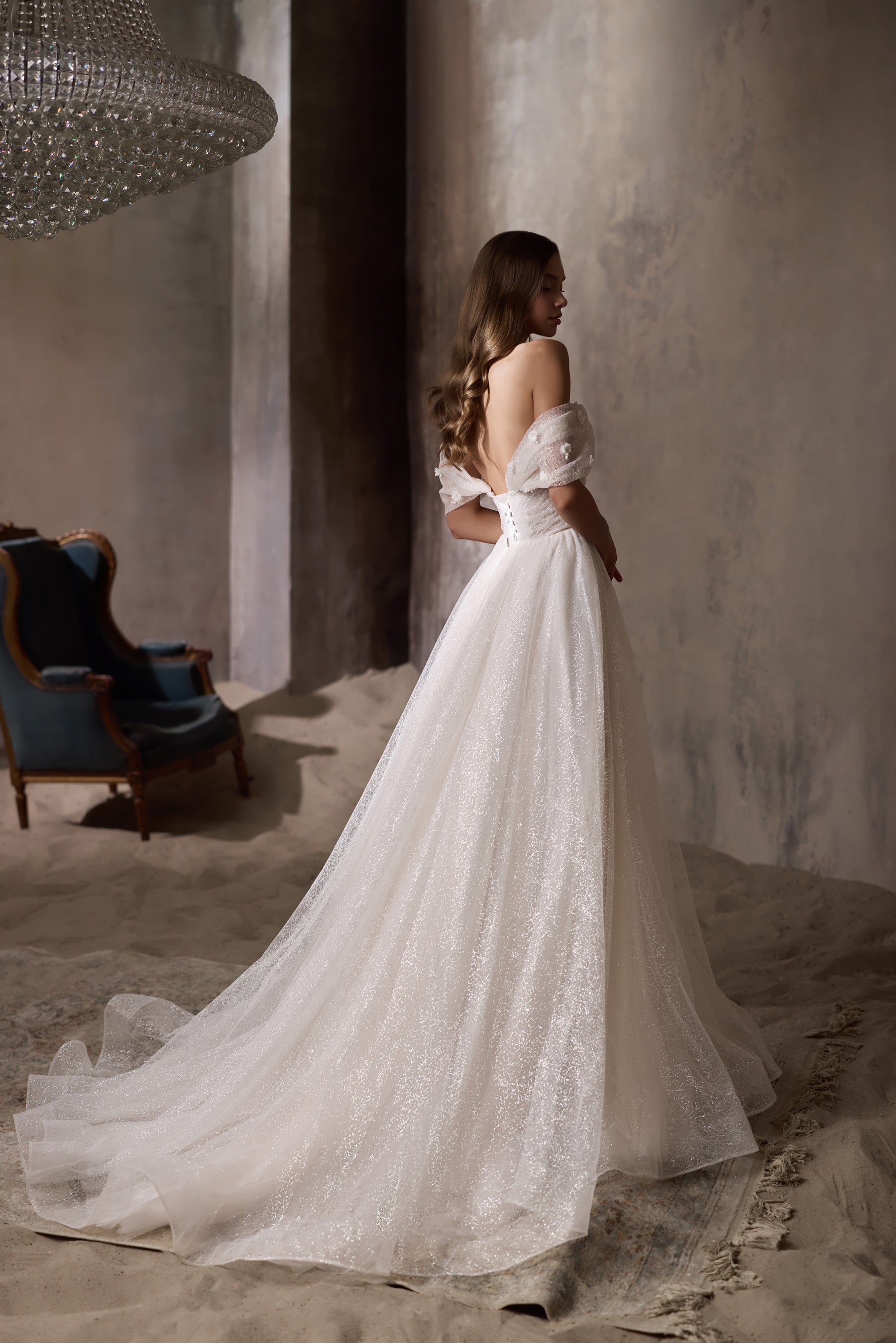 Back view of an ivory wedding dress with an open back, off-the-shoulder sleeves, glittering skirt, and train