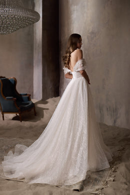 Back view of an ivory wedding dress with an open back, off-the-shoulder sleeves, glittering skirt, and train