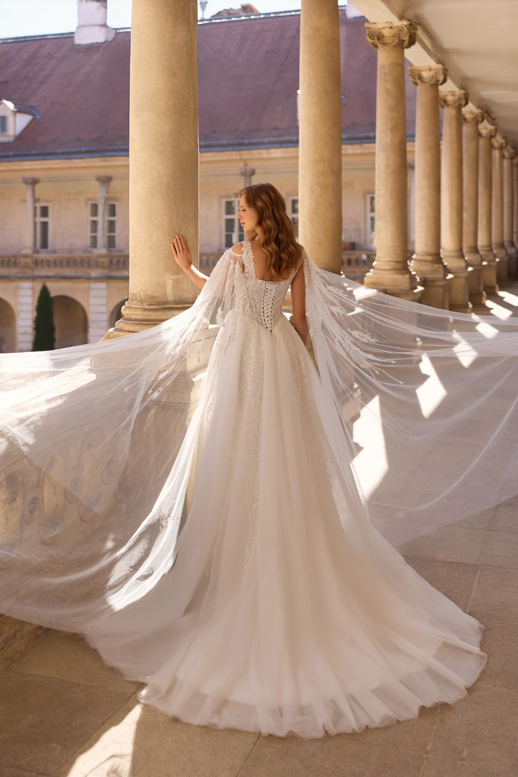 Ivory wedding dress with a flowing A-line skirt, sweeping train, and long sheer cape sleeves shown from behind
