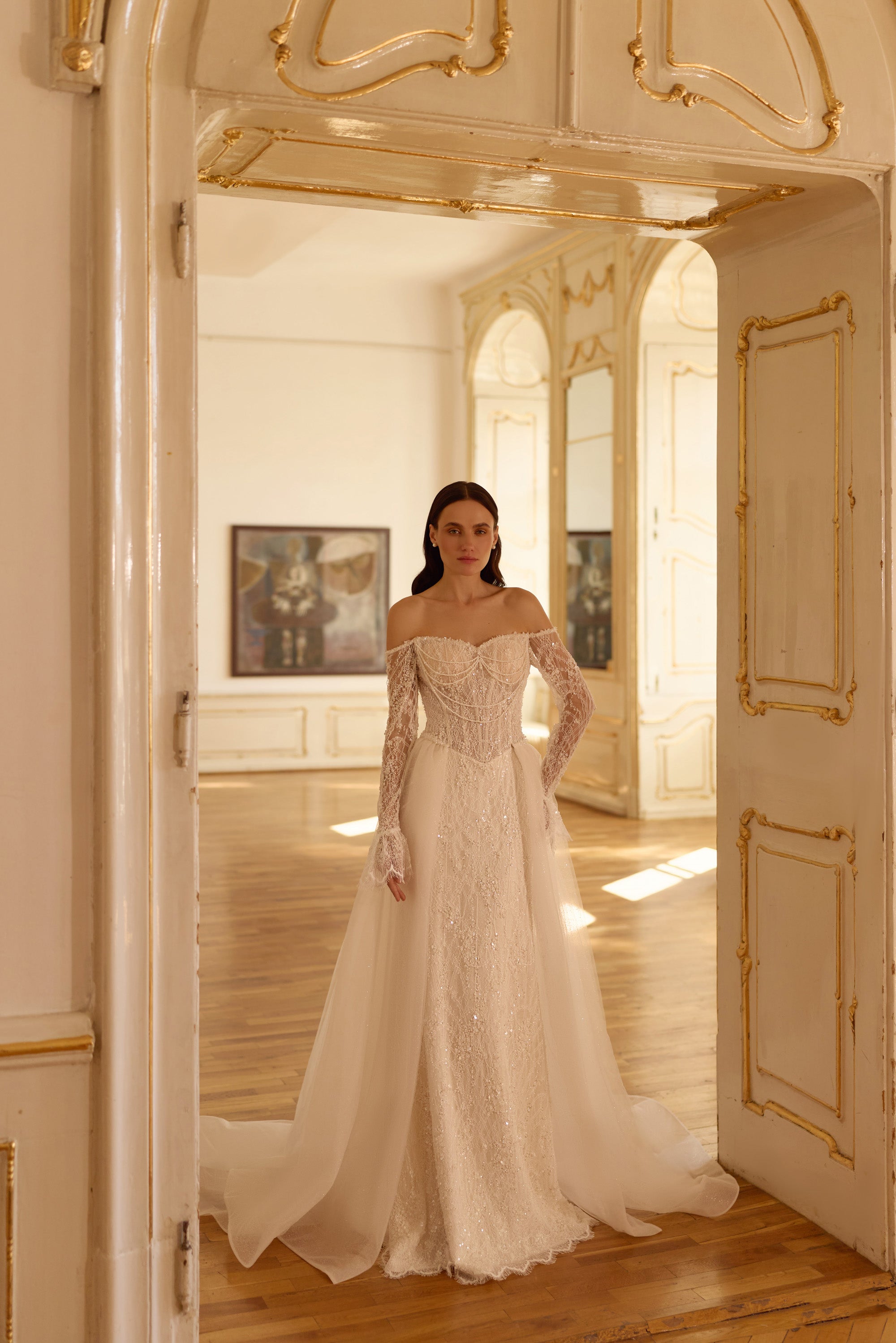 Sparkling ivory wedding dress with an off-shoulder neckline, lace sleeves, fitted skirt, and flowing overskirt