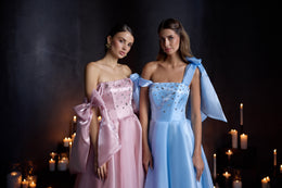 Pink evening dress with a jeweled strapless bodice and sculptural bow drapes, shown beside a blue version
