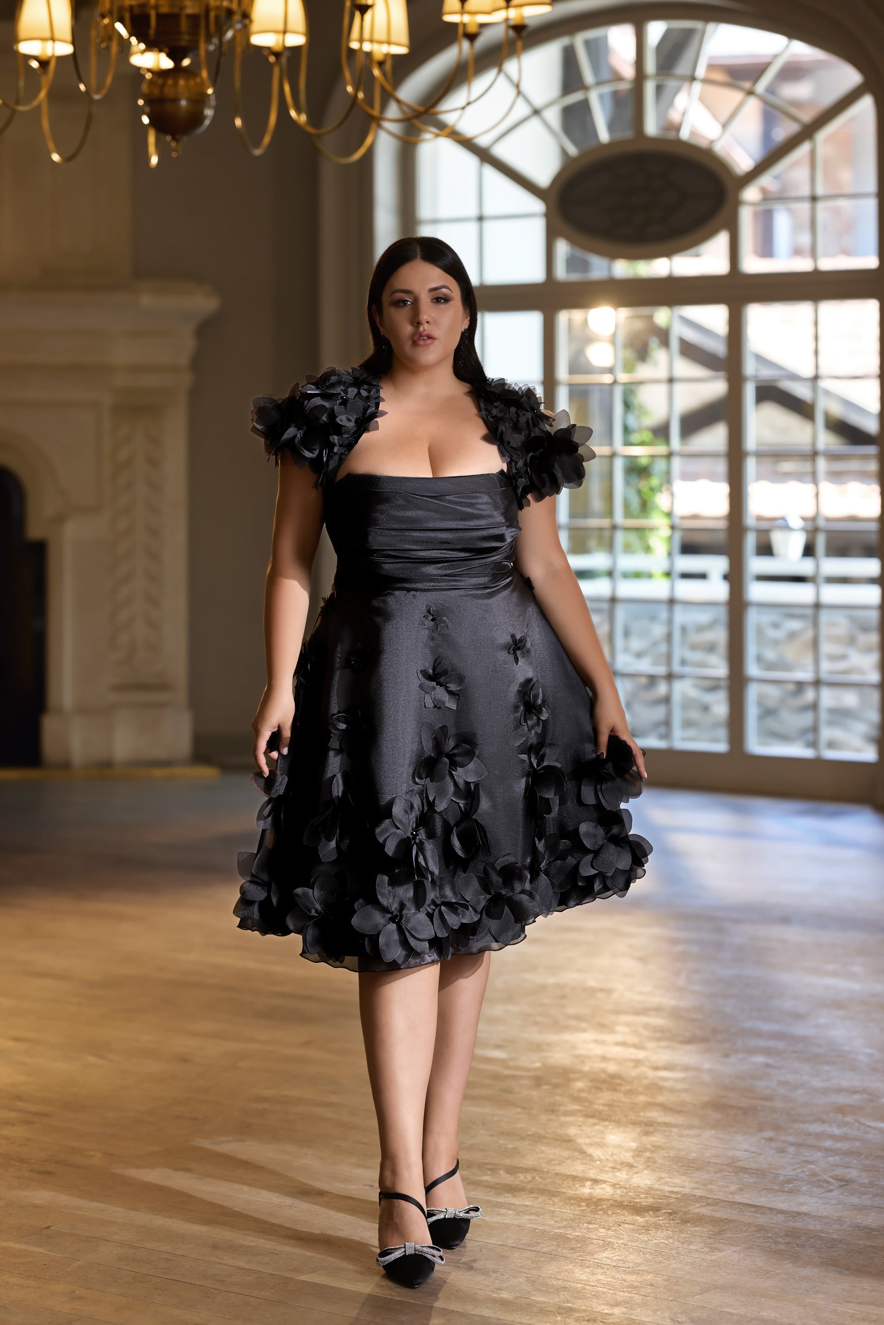 Black knee-length evening dress with a ruched bodice, floral off-shoulder sleeves, and textured appliqués