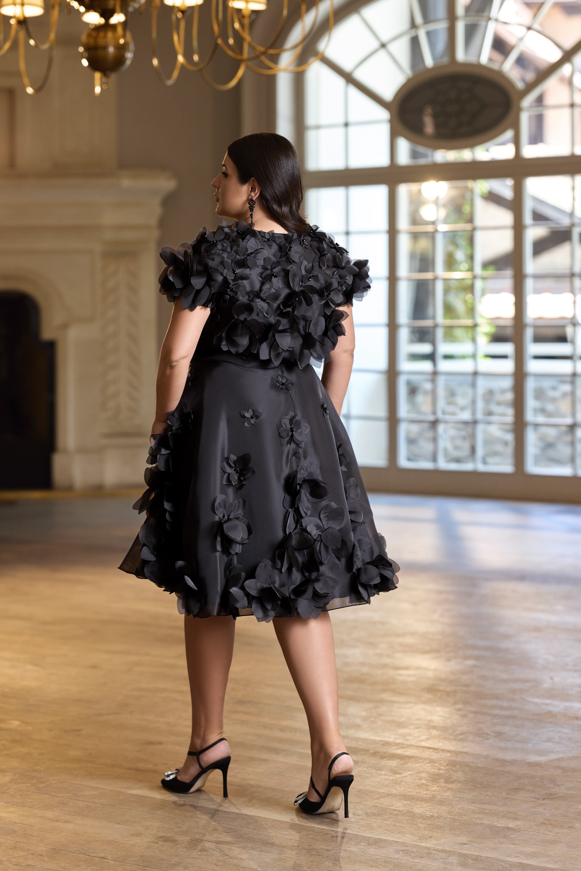 Back of a black evening dress with a floral shoulder overlay, appliquéd skirt, and short flared silhouette