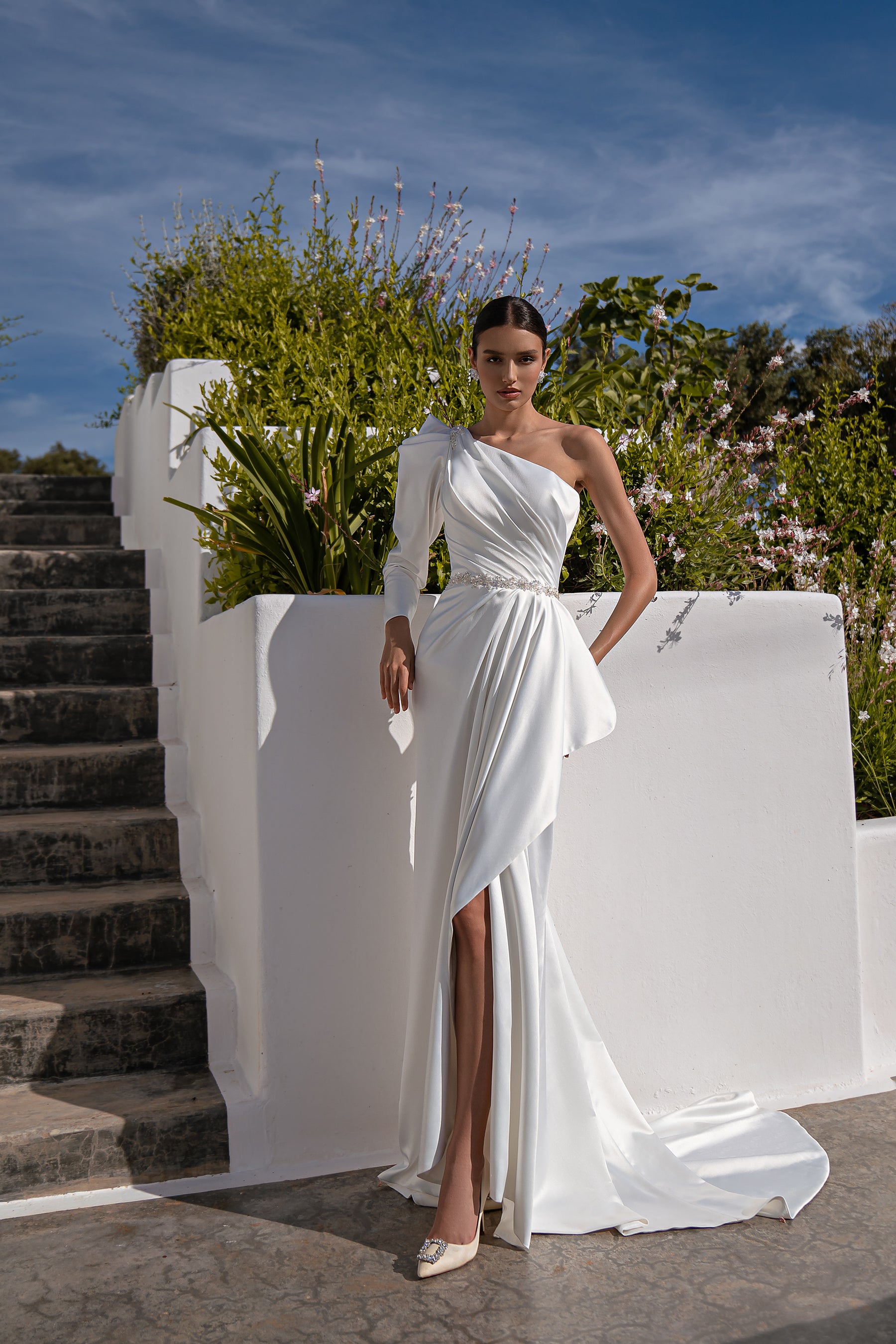 White one-shoulder wedding dress with a beaded waist, thigh-high slit, draped skirt, and train