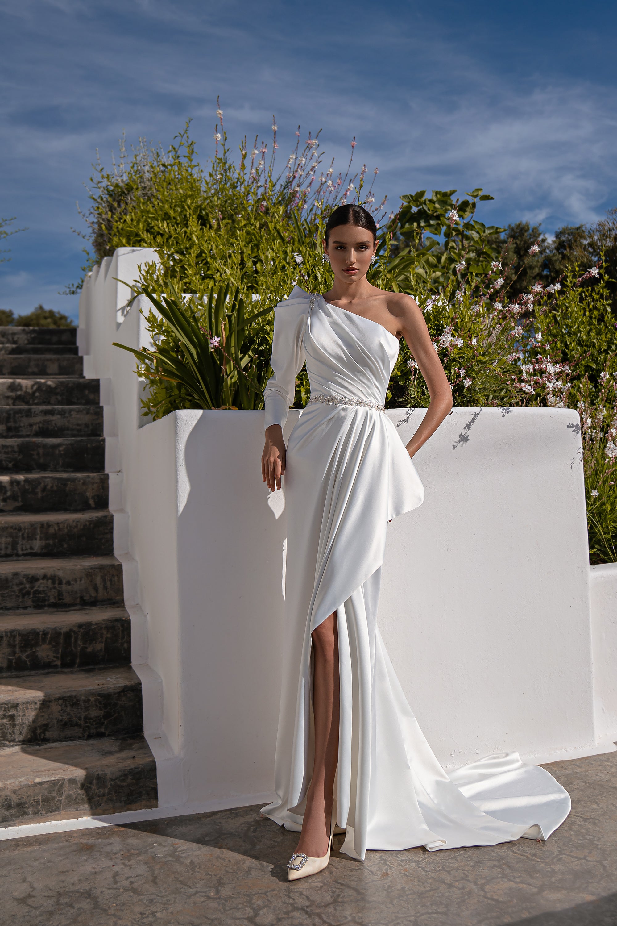 White one-shoulder wedding dress with a beaded waist, thigh-high slit, draped skirt, and train