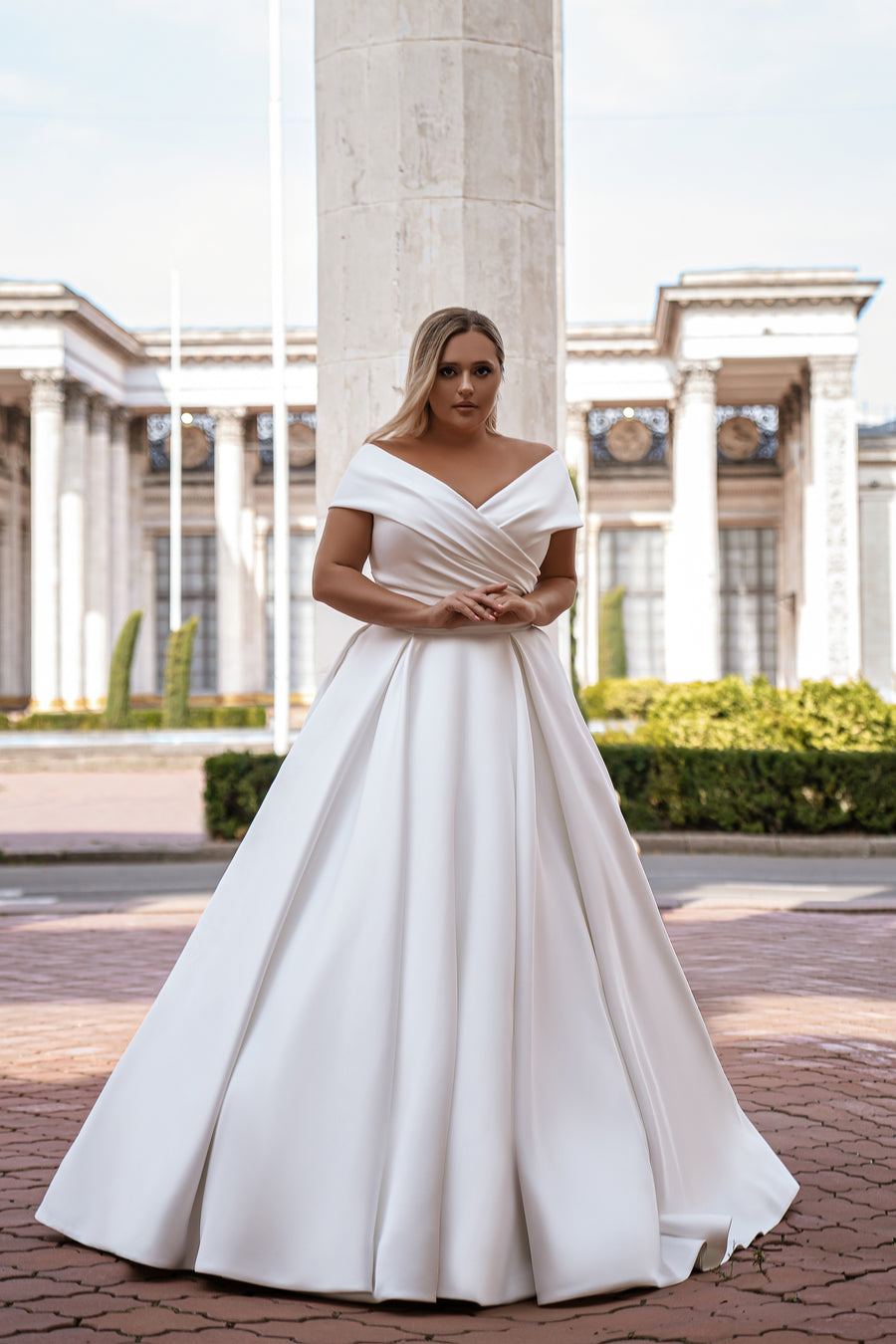 White ball gown wedding dress with a draped off-the-shoulder neckline, fitted waist, and full pleated skirt