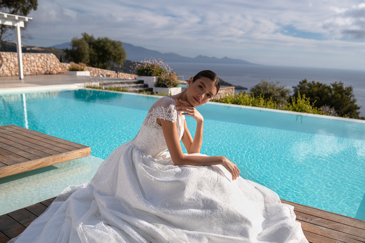 Seated white wedding dress showing short lace sleeves, a sparkling bodice, and a voluminous skirt