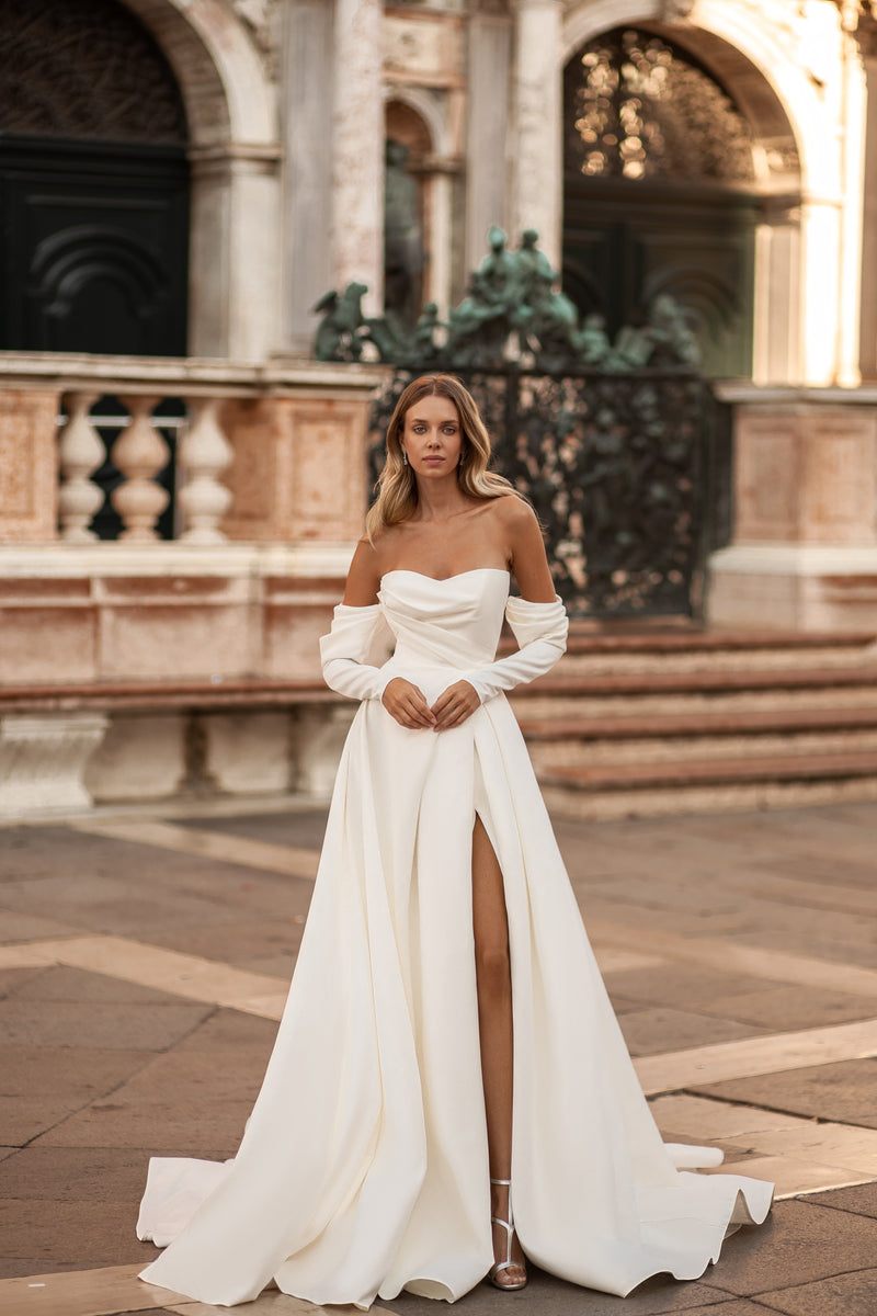 White wedding dress with a draped sweetheart neckline, off-shoulder long sleeves, thigh-high slit, and flowing train