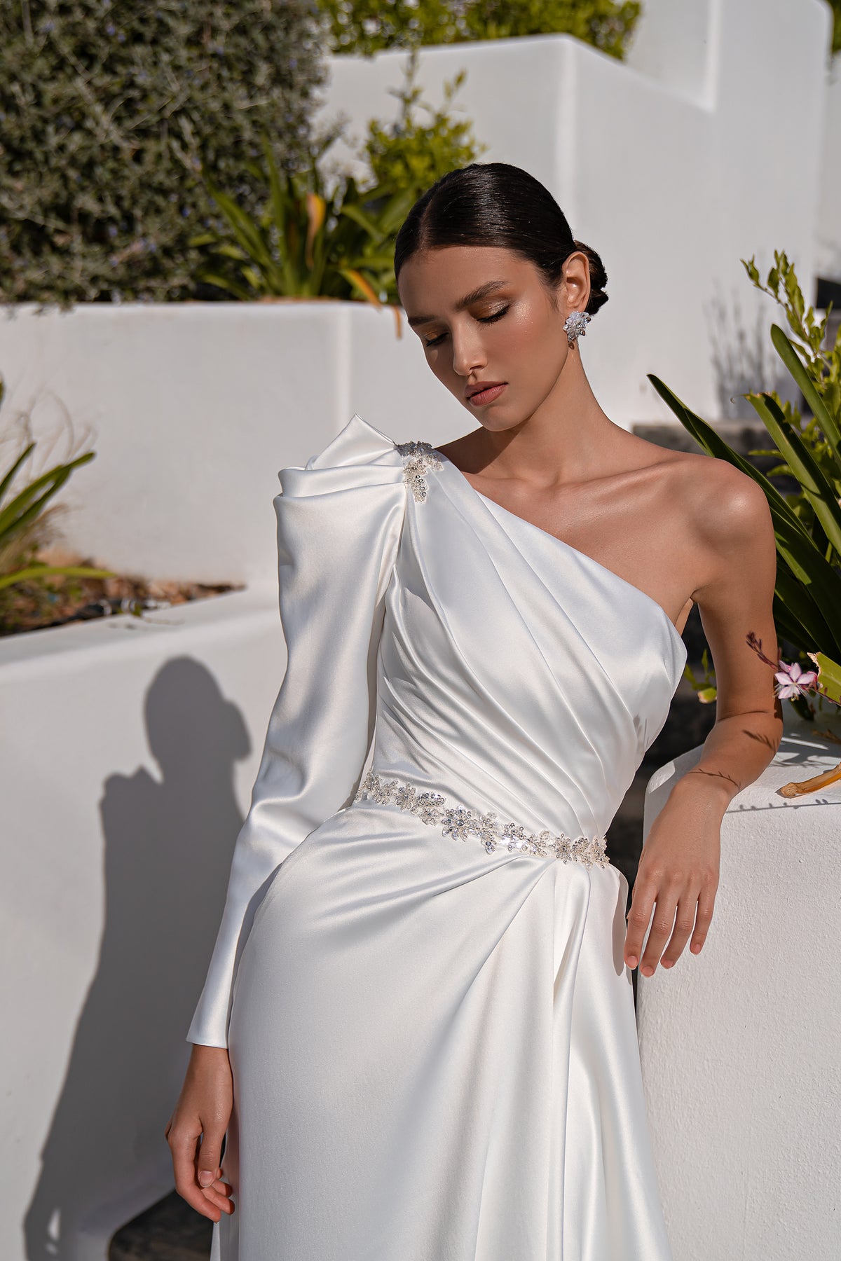 Close-up of a white one-shoulder wedding dress with a long sleeve, draped satin, and beaded waist