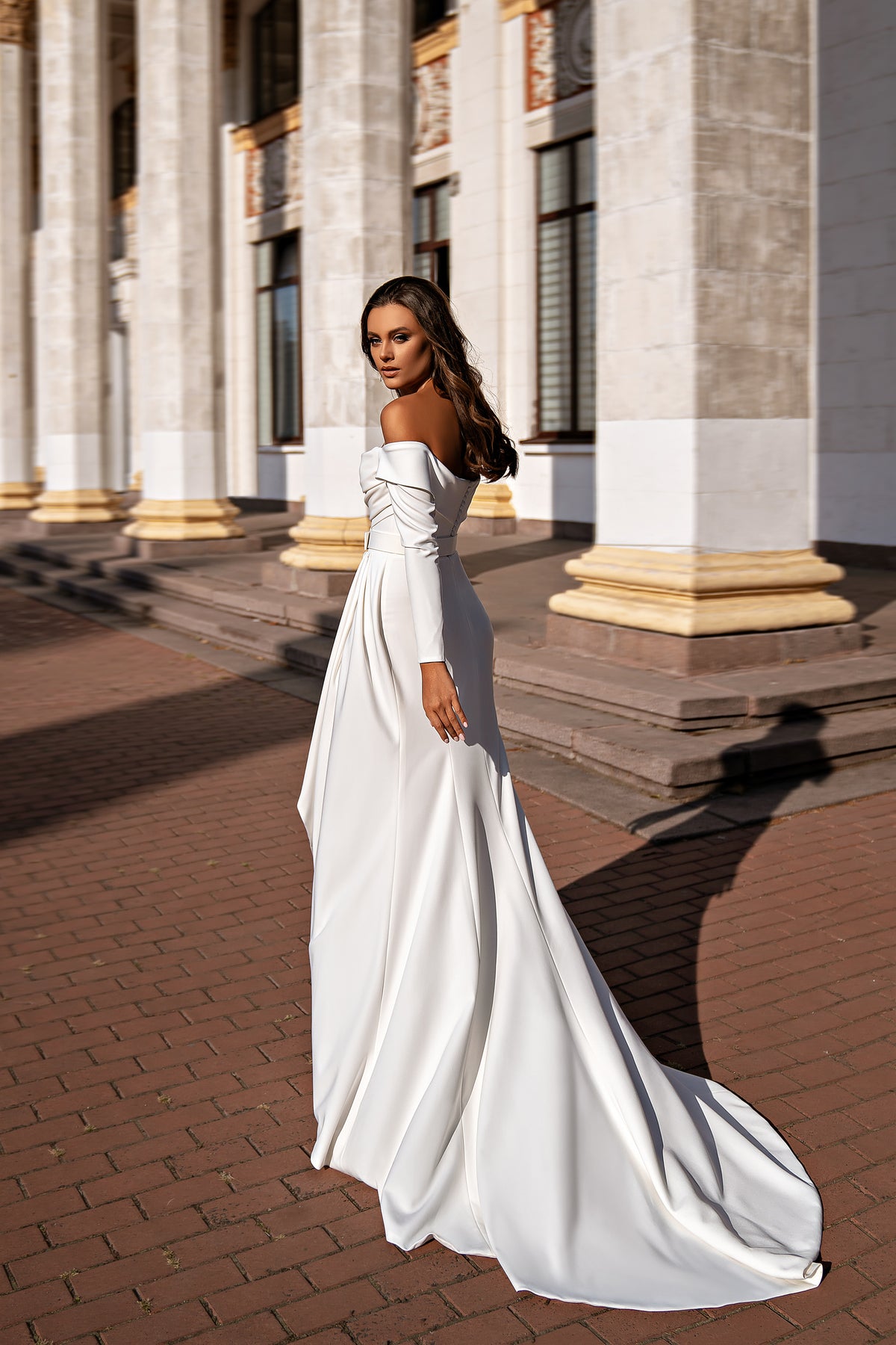 White wedding dress shown from the side with an off-the-shoulder neckline, long sleeves, fitted skirt, and train