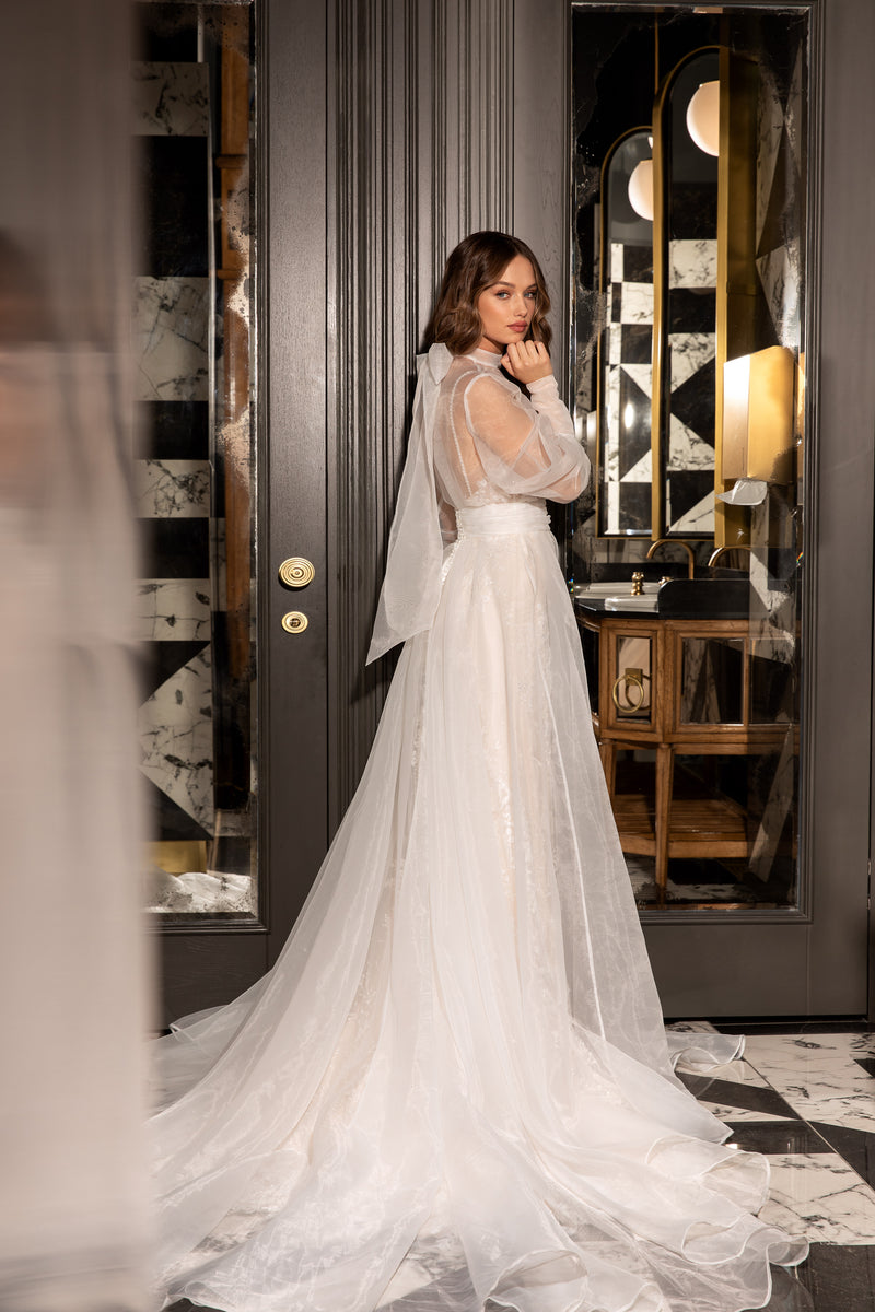 Ivory wedding dress shown with a sheer detachable cape, long sleeves, and a flowing floor-length train