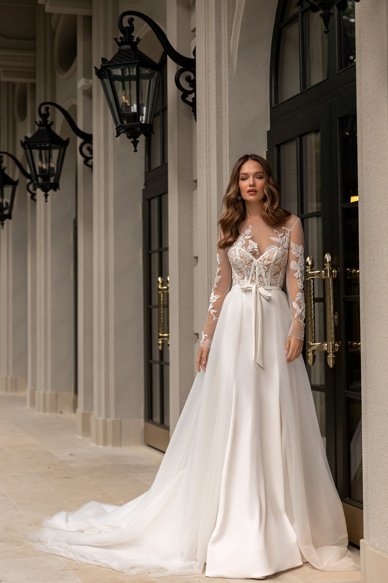 Ivory wedding dress with floral lace bodice, sheer long sleeves, waist bow, and flowing train